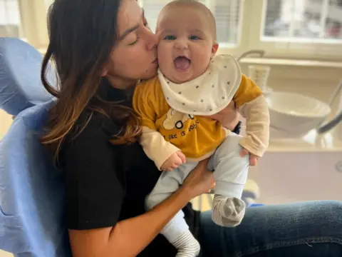 Baş hekimimiz Dr. Ayşenur Kayımbaşıoğlu, AuraDent Klinik odamızda yeni doğmuş bir bebeği muayene etmeden önce elinde tutarken birlikte gülümsüyorlar.